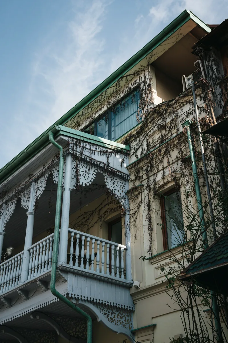 Carved timber balcony with weathered blue details and climbing vines in Tbilisi